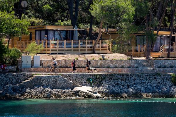 Le persone camminano in una giornata di sole lungo un sentiero accanto a un muro di pietra e case di legno vicino all'acqua.