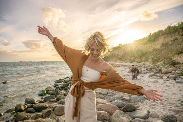 Frau am Strand bei Sonnenuntergang, lächelt und Arme ausgestreckt.
