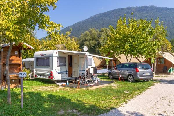Campingplads med en campingvogn og en bil parkeret foran træhuse med bjerge i baggrunden.