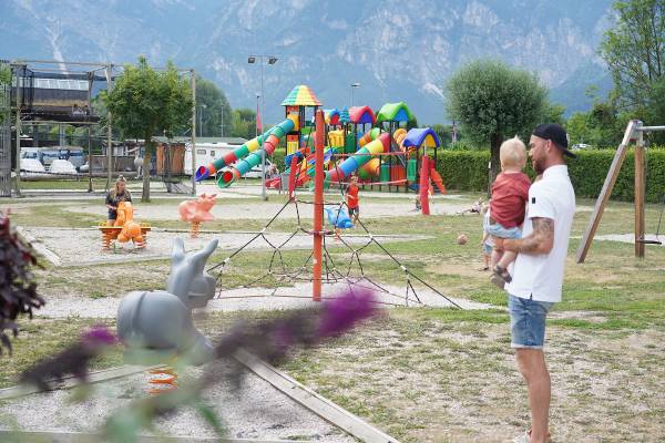 Campingplads med legeplads og familier, bjerge i baggrunden.