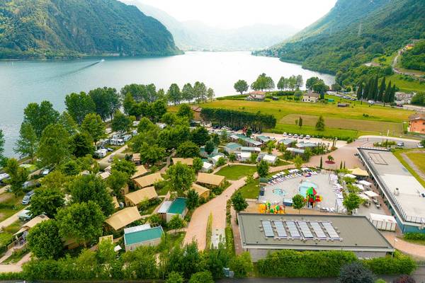 Vue aérienne du camping de glamping de luxe Lago Idro avec un lac, des montagnes et des champs verts.