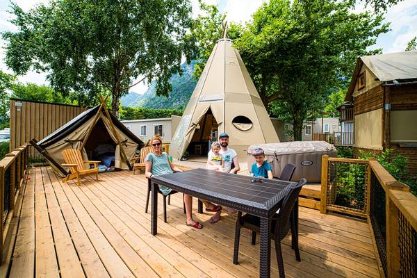 Famille assise à une table sur une terrasse en bois devant un tipi au camping.