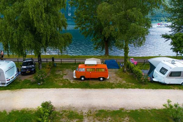 Vue aérienne d'un camping au bord du lac avec un camping-car orange et blanc, des caravanes blanches, des tentes et un bateau au loin.