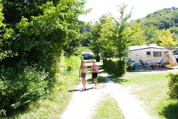Twee meisjes lopen op een campingpad met bomen en geparkeerde campers op de achtergrond.