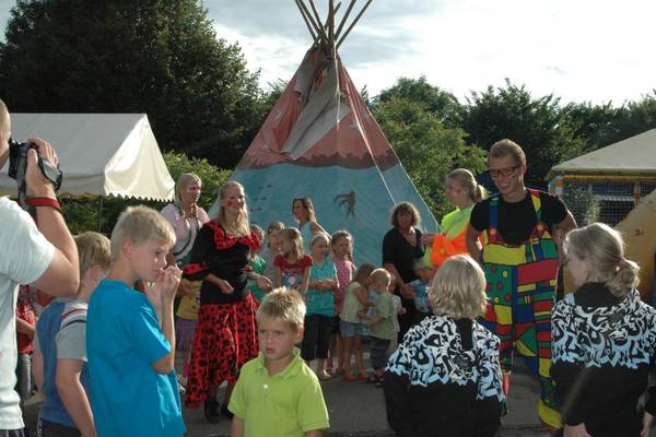 Een groep mensen op een camping, met een geschilderde tipi en een clown in kleurrijke kostuums.