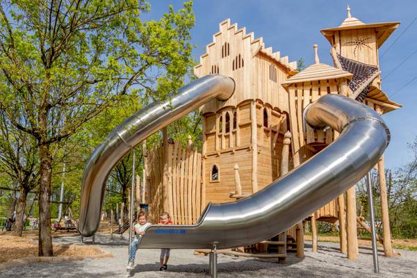 Zwei Kinder stehen an der Basis einer großen Rutsche in einem holzverkleideten Burgspielplatz.
