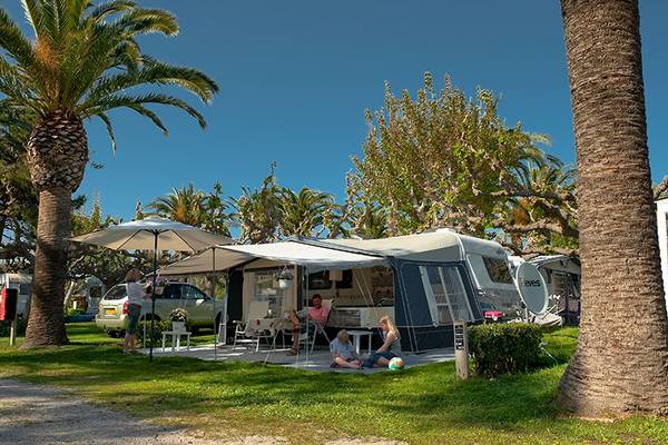 Familie slapper af under en telt på Playa Montroig Camping Resort med palmer og en bil i baggrunden.