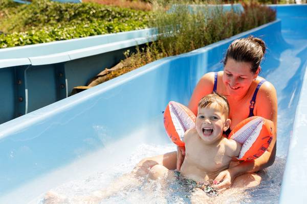 Eine Frau und ein Kind mit Schwimmflügeln gleiten an Camping De Paal eine blaue Wasserrutsche hinunter.