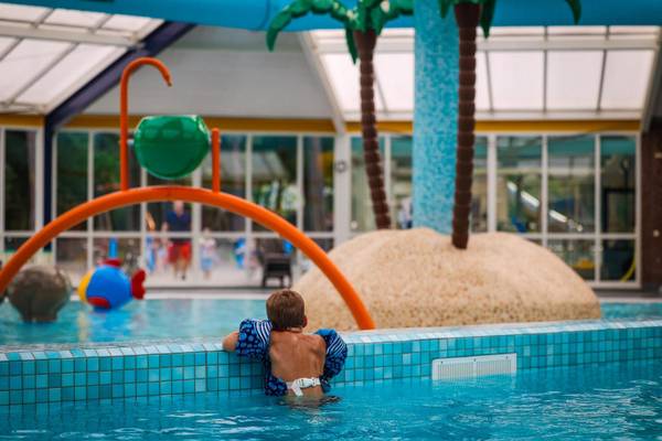 Kind im Schwimmbad im Camping De Paal mit blauen Fliesen und künstlichen Palmen.