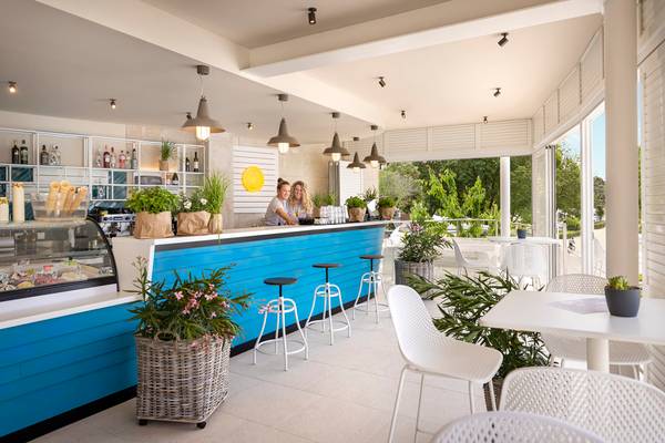 Deux femmes debout derrière un comptoir de bar bleu avec des tabourets et des plantes sur une terrasse au Camping Vestar.
