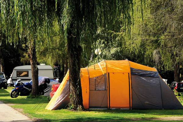 Oranges Zelt auf dem Campingplatz Ferienparadies Natterer See mit Bäumen und geparkten Fahrzeugen im Hintergrund.