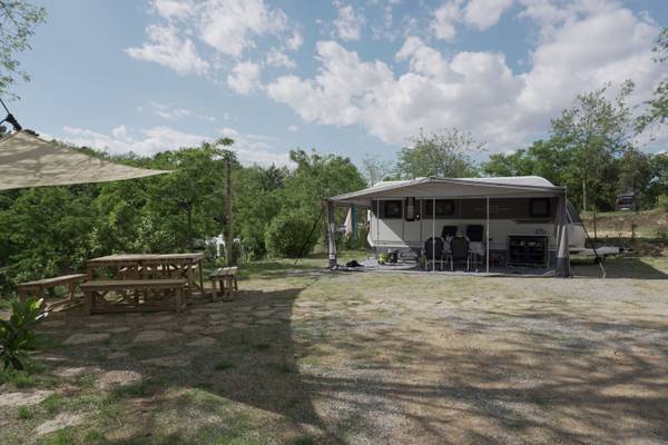 Campingplatz mit Wohnwagen unter Zelt umgeben von Bäumen und Bänken.