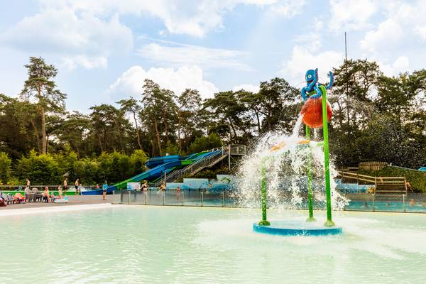 Wasserspielplatz im Camping De Paal mit einer Fontäne in der Mitte und Bäumen im Hintergrund.