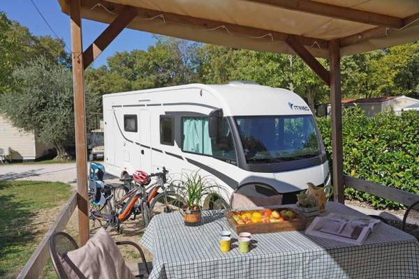 Campingplads med autocamper og cykler parkeret under en trægavl.