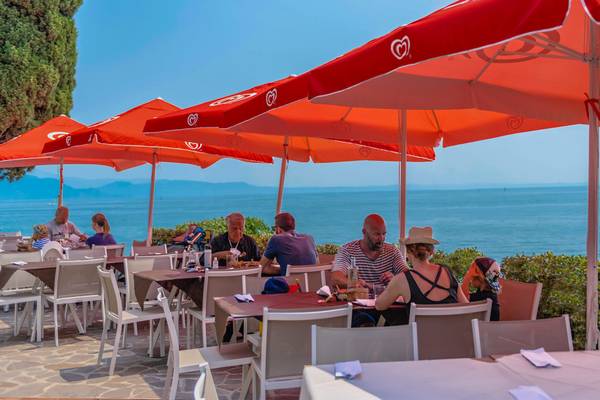 Mennesker sidder ved borde på terrassen i Fornella Camping & Wellness Family Resort med røde parasoller med udsigt til havet.