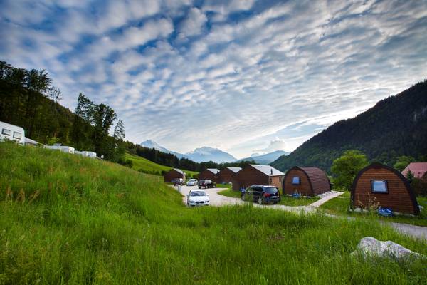 Campingplatz mit Holzhütten, geparkten Autos und einer Berglandschaft im Hintergrund.