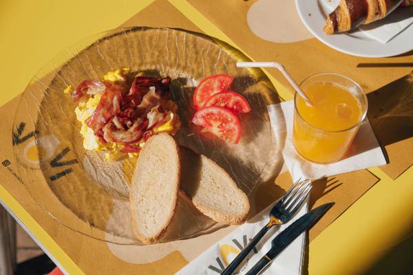 Plato de desayuno con tocino, tomates, pan y jugo de naranja sobre una mesa amarilla.