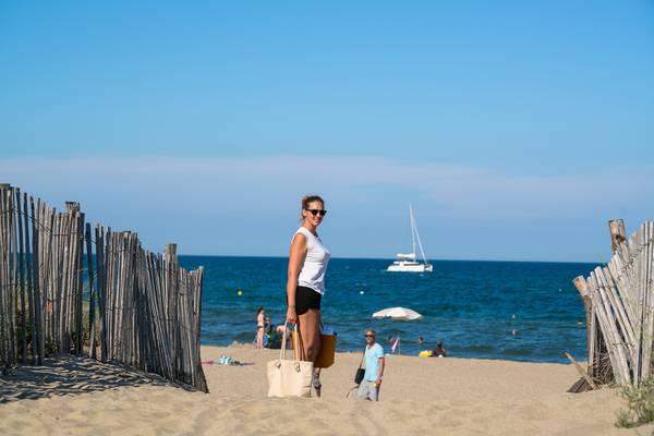 Femme à la plage avec lunettes de soleil et sacs, océan et voilier en arrière-plan.