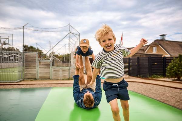 Two children playing on a trampoline at Henne Strand Resort, with a man upside down and a flag in the background.