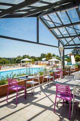 Terrasse du Camping Le Pin Parasol avec des tables et des chaises et une piscine en arrière-plan.