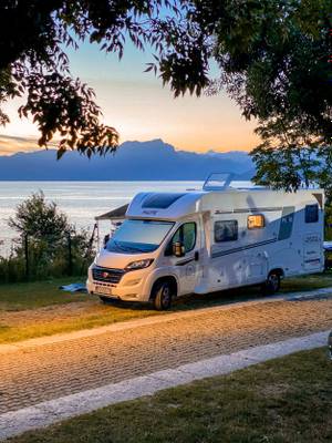 En hvid campingvogn parkeret under et træ ved en sø med bjerge i baggrunden.
