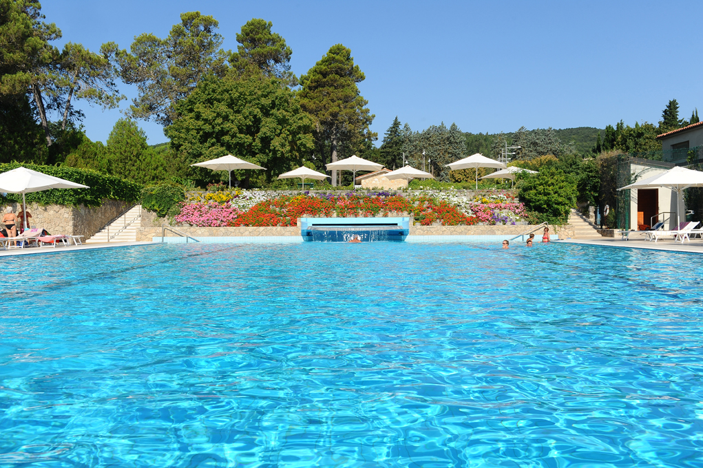 Swimming pool with blue water, surrounded by umbrellas, flowers, and people.
