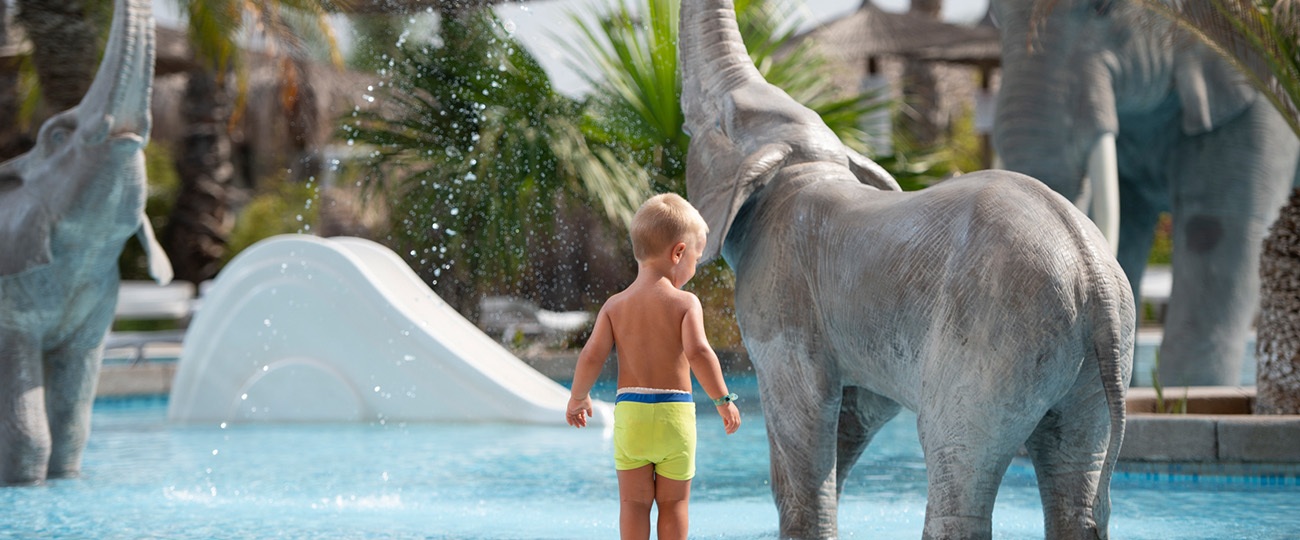 A young boy stands by an elephant statue in a swimming pool.
